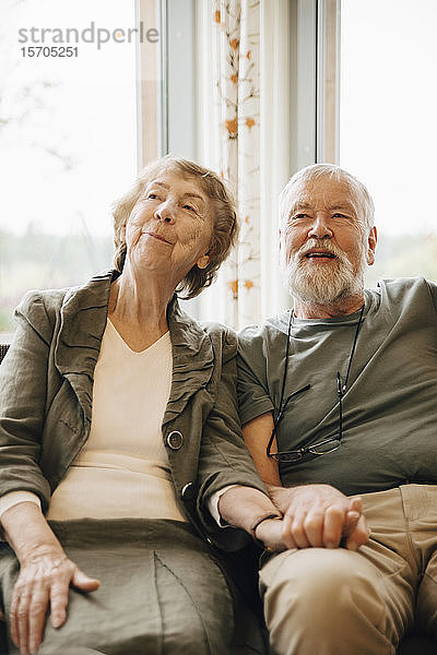 Älteres Ehepaar hält sich an den Händen  während es am Fenster eines Pflegeheims sitzt