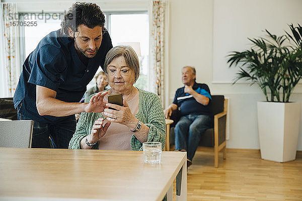 Zuversichtlicher männlicher Pfleger unterstützt ältere Frau bei der Nutzung eines Smartphones im Pflegeheim