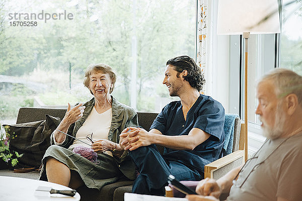 Lächelnde ältere Frau im Gespräch mit einem Krankenpfleger  der auf einem Sofa vor dem Fenster eines Pflegeheims sitzt