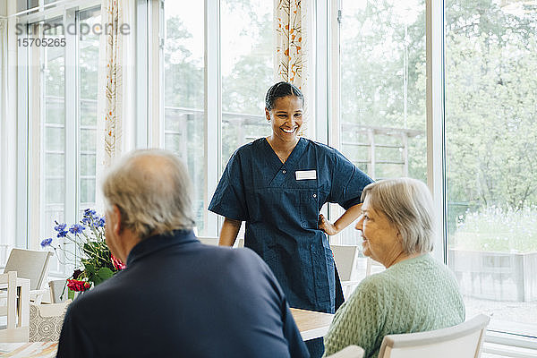 Lächelnde junge Krankenschwester sieht ältere Frau an  die von einem Mann am Esstisch im Altenpflegeheim sitzt