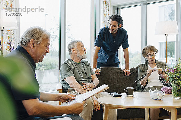 Männlicher Mitarbeiter im Gesundheitswesen im Gespräch mit einem älteren Mann  der auf einem Sofa inmitten von Freunden im Altenheim sitzt
