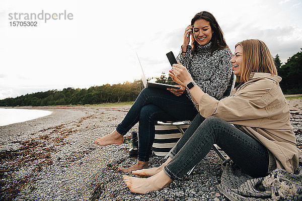 Glückliche Freunde schauen auf Smartphones  während sie am Strand am Ufer gegen den Himmel sitzen