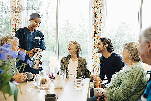 Lächelnde Krankenschwester serviert einer älteren Frau  die mit Freunden und Enkel am Esstisch im Pflegeheim sitzt  ein Getränk