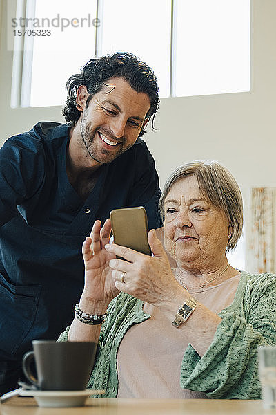 Lächelnder männlicher Betreuer unterstützt ältere Frau bei der Nutzung eines Smartphones im Pflegeheim