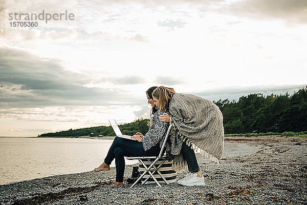 Freundin begrüßt Frau  die am Laptop arbeitet  während sie am Strand gegen den Himmel sitzt