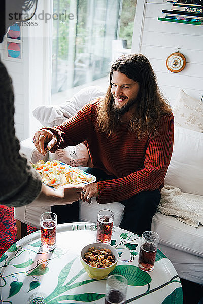 Schrägaufnahme eines Freundes  der einem Mann Snacks serviert  während er zu Hause auf dem Sofa sitzt