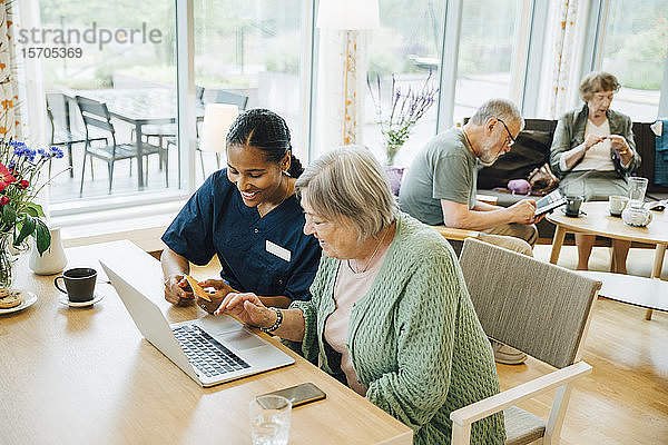 Lächelnde Gesundheitshelferin unterstützt ältere Frau beim Online-Shopping mit Kreditkarte und Laptop im Pflegeheim