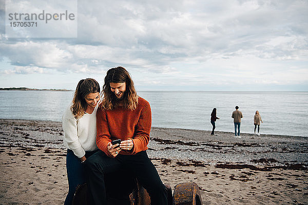 Ehepaar  das am Strand am Meer sitzend auf sein Handy schaut