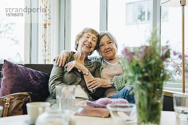Lächelnde ältere Frauen sitzen mit umarmtem Arm auf dem Sofa vor dem Fenster im Altersheim