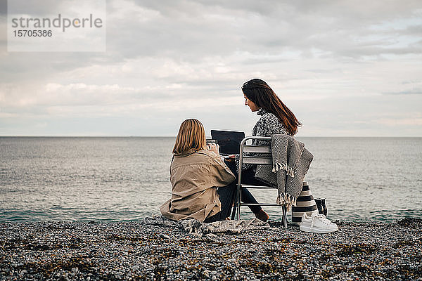 Freundinnen  die Technologien nutzen  während sie am Strand vor bewölktem Himmel sitzen