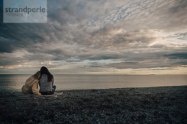 Rückansicht von liebenden Freunden  die am Meeresufer am Strand vor bewölktem Himmel sitzen