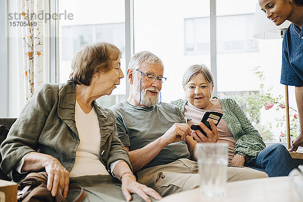 Älterer Mann benutzt Smartphone  während er inmitten von Frauen im Altersheim sitzt
