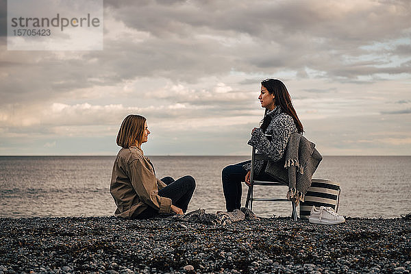 Freundinnen unterhalten sich  während sie am Strand vor bewölktem Himmel sitzen