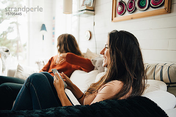 Lächelnde Frau mit Smartphone  die wegschaut  während sie mit ihrem Freund zu Hause auf dem Sofa sitzt