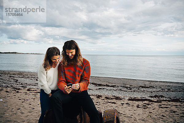 Lächelnder Mann zeigt der Frau sein Handy  während er am Strand sitzt