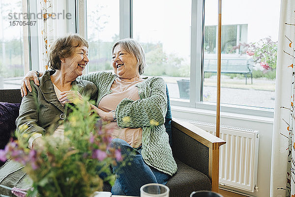 Lächelnde ältere Frauen sitzen mit umarmtem Arm und schauen sich auf dem Sofa gegen das Fenster im Altersheim an