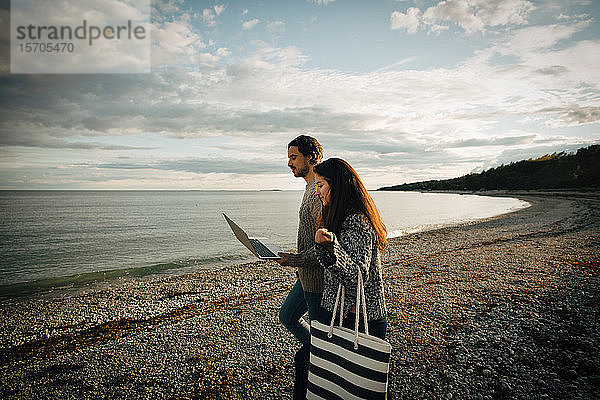 Frau geht mit dem Mann mit Laptop am Strand am Meeresufer gegen den Himmel