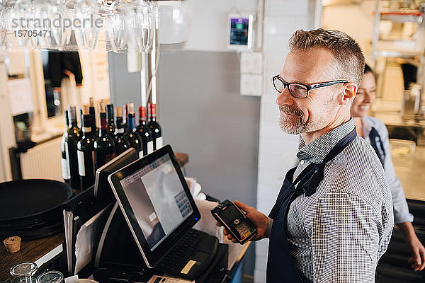 Lächelnder Mann benutzt Computer  während er im Restaurant ein Kreditkartenlesegerät in der Hand hält