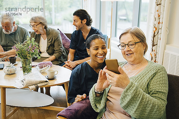 Pensionierte ältere Frau teilt ihr Smartphone mit lächelnder junger Krankenschwester beim Gemeindeeinsatz