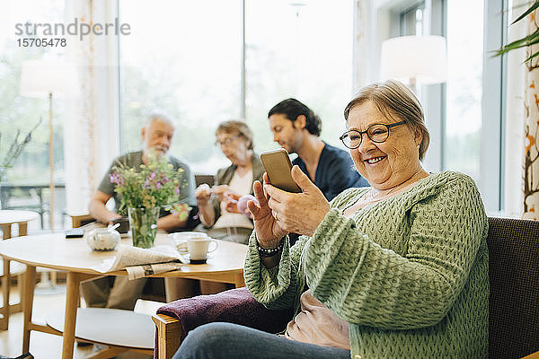 Lächelnde Rentnerin im Ruhestand benutzt Smartphone  während sie mit Freunden und Betreuer im Hintergrund auf einem Stuhl sitzt