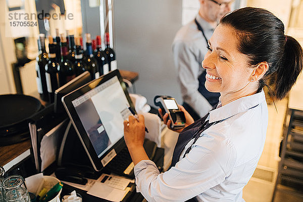 Lächelnde Frau benutzt Computer  während sie im Restaurant ein Kreditkartenlesegerät in der Hand hält