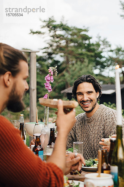 Porträt eines lächelnden Mannes  der mit Freunden am Esstisch bei einer Gartenparty sitzt