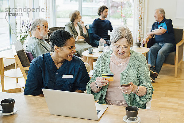 Fröhliche  von einer älteren Frau sitzende Gesundheitshelferin beim Online-Shopping mit Kreditkarte und Smartphone in Krankenschwestern