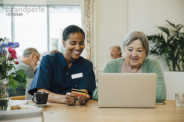 Lächelnde Pflegerin unterstützt ältere Frau beim Online-Shopping mit Kreditkarte und Laptop im Pflegeheim