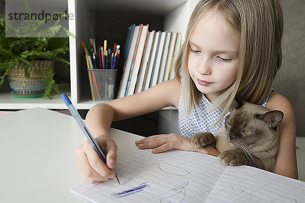 Mädchen mit einer Katze  die zu Hause am Tisch sitzt und Hausaufgaben macht
