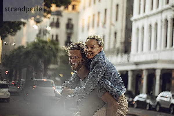 Glücklicher junger Mann trägt seine Freundin huckepack durch die Stadt