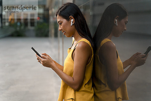 Modisch gekleidete Frau in Gelb lehnt mit Kopfhörern und Smartphone an Glasfront