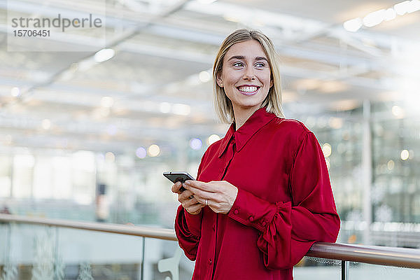 Lächelnde junge Geschäftsfrau steht mit Handy an einem Geländer