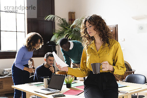Frau mit Papier und Kaffeetasse im Büro mit Kollegen im Hintergrund