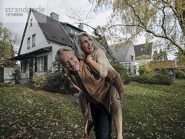 Glücklicher älterer Mann  der seine Frau im Herbst huckepack im Garten seines Hauses trägt