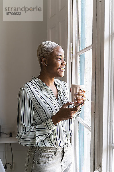 Glückliche Frau mit Handy und Kaffeetasse  die aus dem Fenster schaut