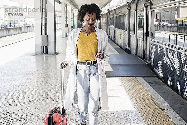 Lächelnde junge Frau mit Kopfhörern und Smartphone am Bahnsteig