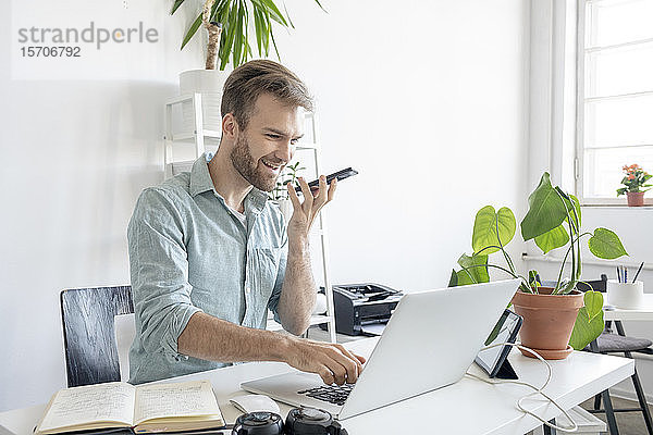 Lächelnder Mann mit Smartphone und Laptop am Schreibtisch im Büro