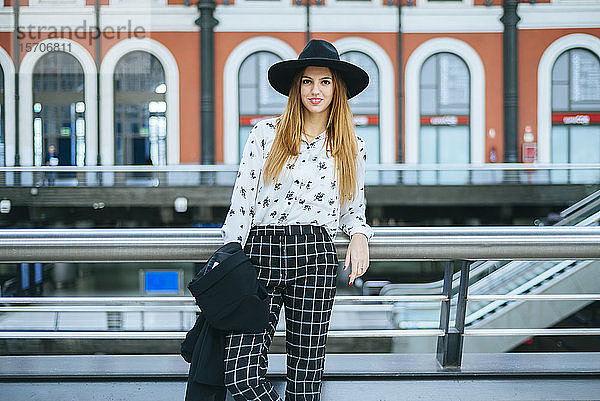 Porträt einer lächelnden jungen Frau mit Hut am Bahnhof