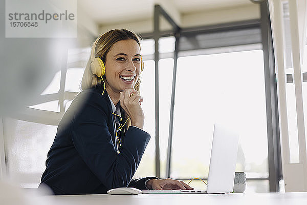 Porträt einer glücklichen jungen Geschäftsfrau mit Kopfhörer und Laptop am Schreibtisch im Büro