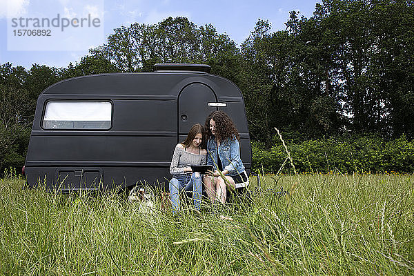 Zwei beste Freunde sitzen auf einer Wiese vor schwarzem Wohnwagen mit digitalem Tablet