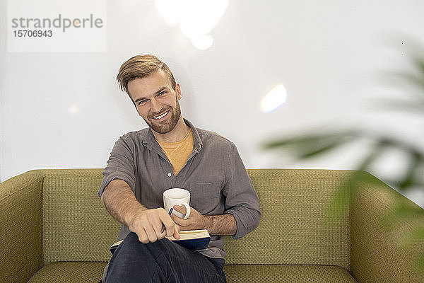 Porträt eines lächelnden Mannes auf einer Couch sitzend mit Buch und Kaffeetasse
