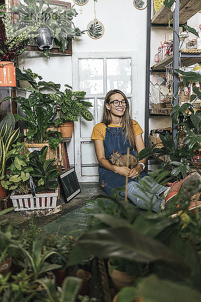 Glückliche junge Frau mit Dinosaurier-Figur  die in einem kleinen Laden mit Pflanzen auf dem Boden sitzt
