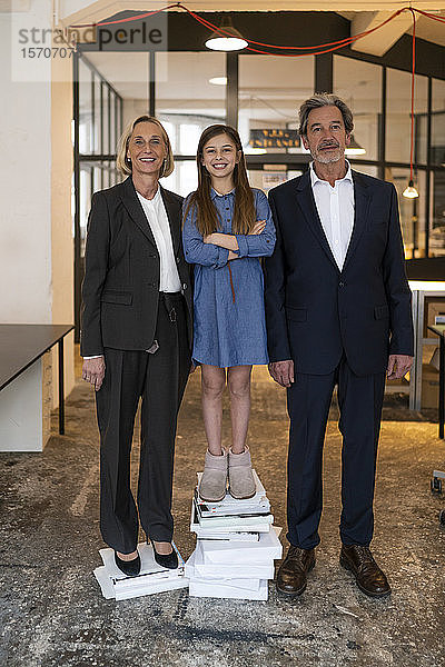 Porträt eines lächelnden Geschäftsmannes und einer lächelnden Geschäftsfrau mit Mädchen auf einem Stapel Bücher im Büro stehend
