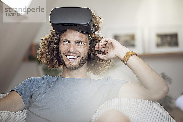 Porträt eines lächelnden jungen Mannes  der zu Hause eine VR-Brille trägt