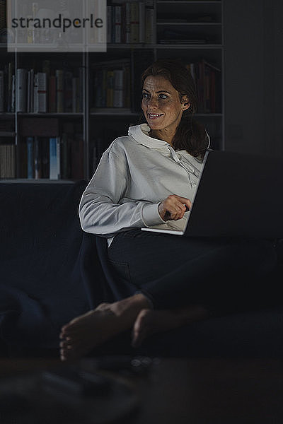 Frau sitzt auf der Couch eines dunklen Wohnzimmers und benutzt einen Laptop
