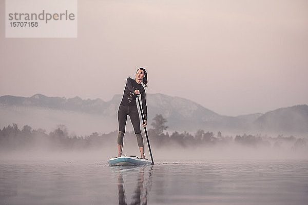 Frau steht auf und paddelt auf dem Kirchsee im Morgennebel  Bad Tölz  Bayern  Deutschland