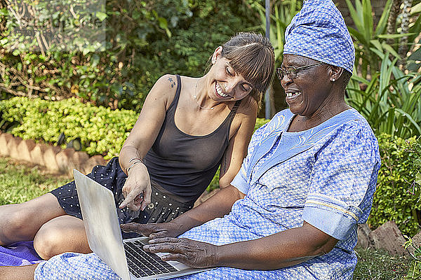 Glückliche ältere Frau sitzt auf dem Rasen und teilt ihren Laptop mit einer Frau