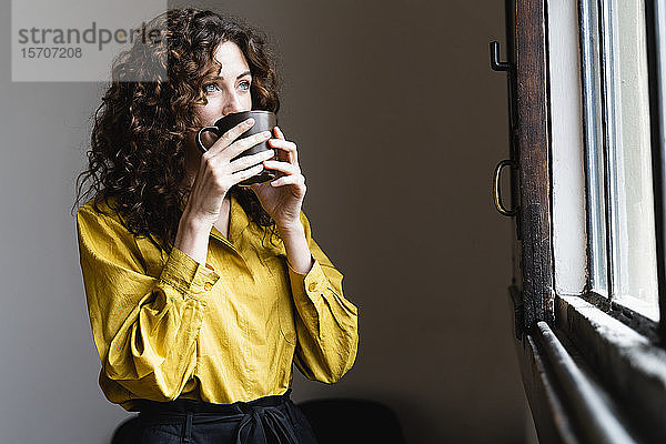 Frau schaut aus dem Fenster und trinkt aus Kaffeetasse