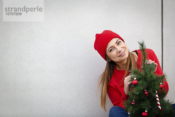 Frau mit rotem Pullover und Wollmütze  die einen künstlichen Weihnachtsbaum vor eine Wand hält