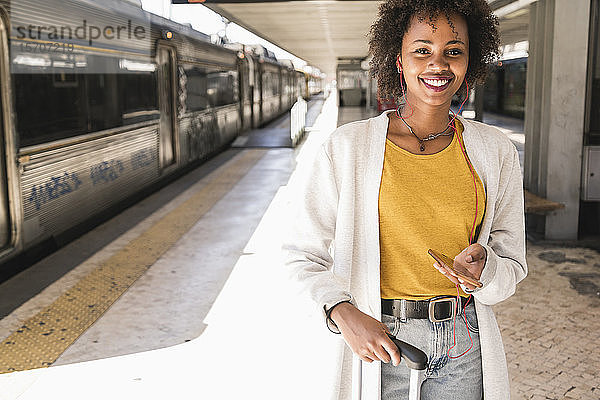 Porträt einer lächelnden jungen Frau mit Kopfhörern und Smartphone am Bahnsteig